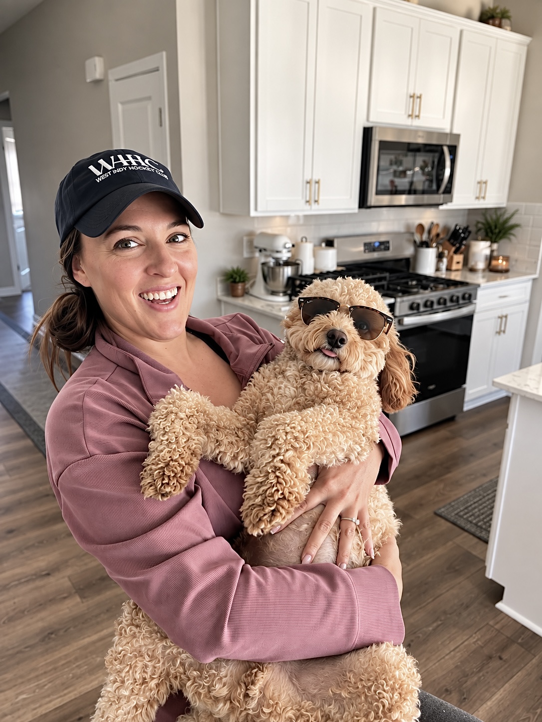 The owner smiling at the camera while holding her curly golden doodle, who is wearing sunglasses
